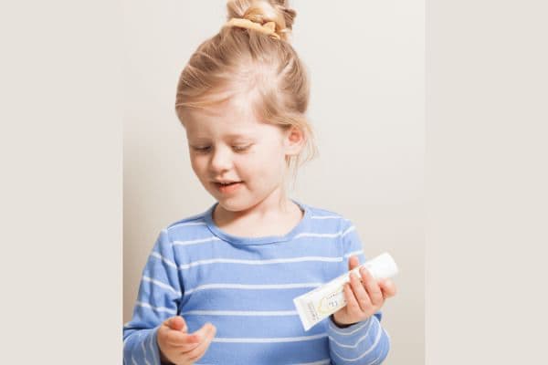 a little blonde girl holding a tube of toothpaste in her left hand, and looking down at her right hand